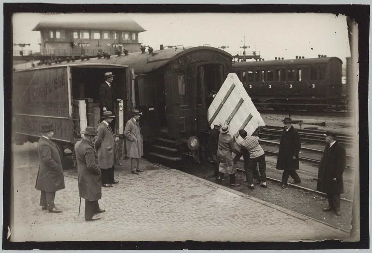 Photo noir et blanc de manutentionnaires hissant une grande caisse en bois dans un wagon de train, sous le regard d’hommes en manteau et chapeau