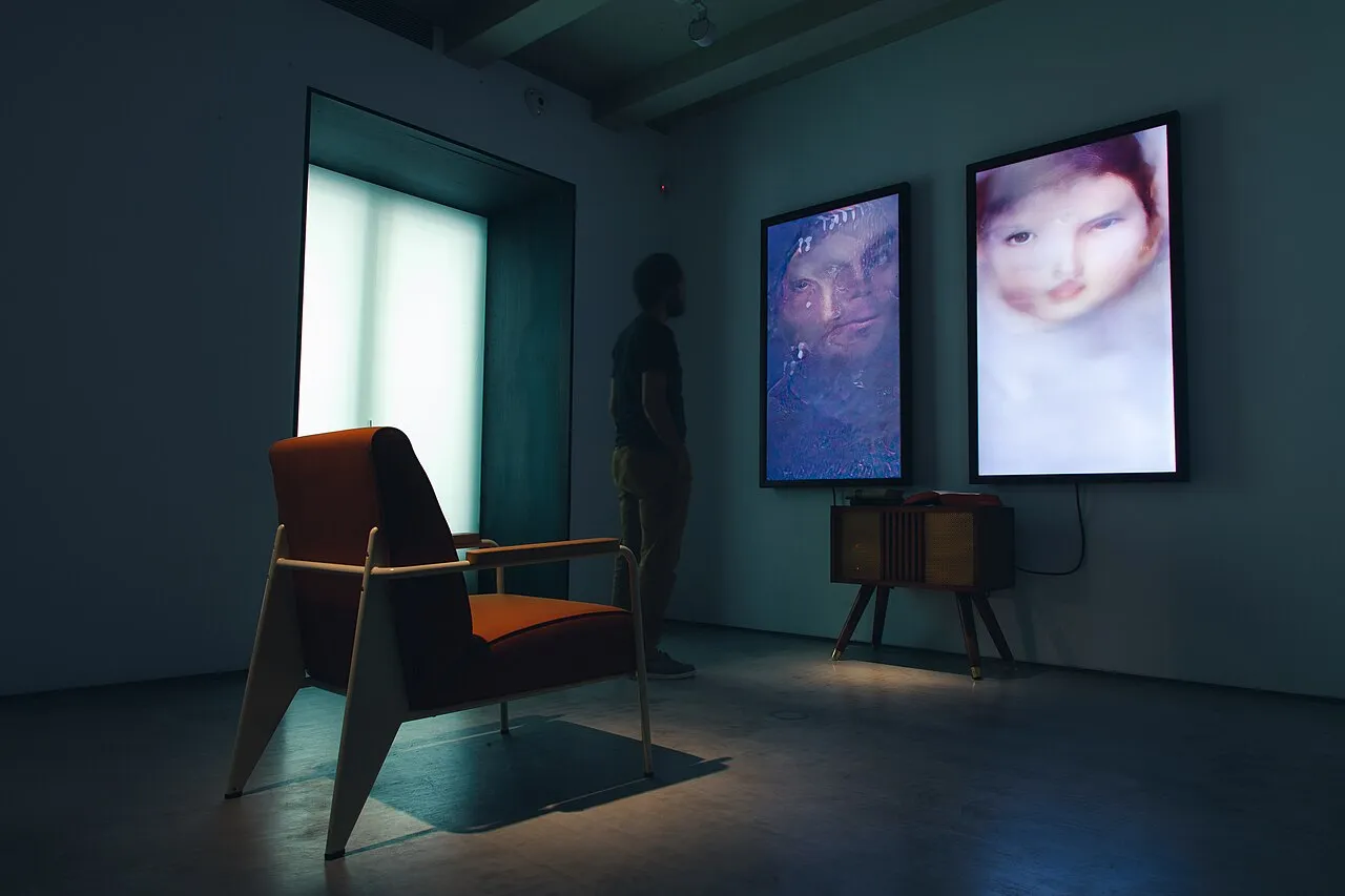 Salle sombre avec deux écrans verticaux affichant des portraits générés par ordinateur au-dessus d’un meuble en bois, un fauteuil au premier plan et un visiteur de dos