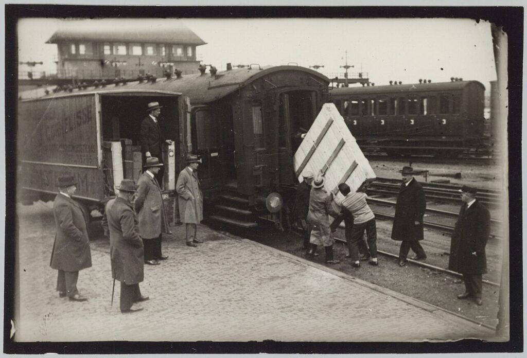 Photo noir et blanc de manutentionnaires hissant une grande caisse en bois dans un wagon de train, sous le regard d'hommes en manteau et chapeau