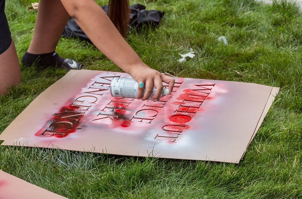 Main tenant une bombe de peinture au-dessus d'un pochoir posé sur un carton, lettres rouges sur l'herbe