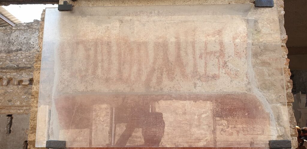 Grandes lettres rouges peintes sur un mur antique de Pompéi, protégées par une vitre