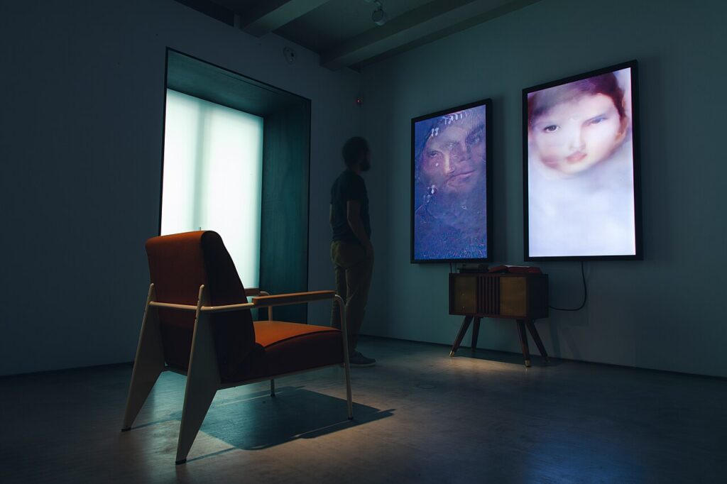 Salle sombre avec deux écrans verticaux affichant des portraits générés par ordinateur au-dessus d'un meuble en bois, un fauteuil au premier plan et un visiteur de dos